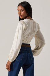 A woman with long, straight black hair is wearing a cream-colored, long-sleeved blouse with a subtle sheen. The blouse has a gathered elastic waist and delicate eyelet lace detail along the neckline. She pairs it with high-waisted dark blue jeans and a brown belt.